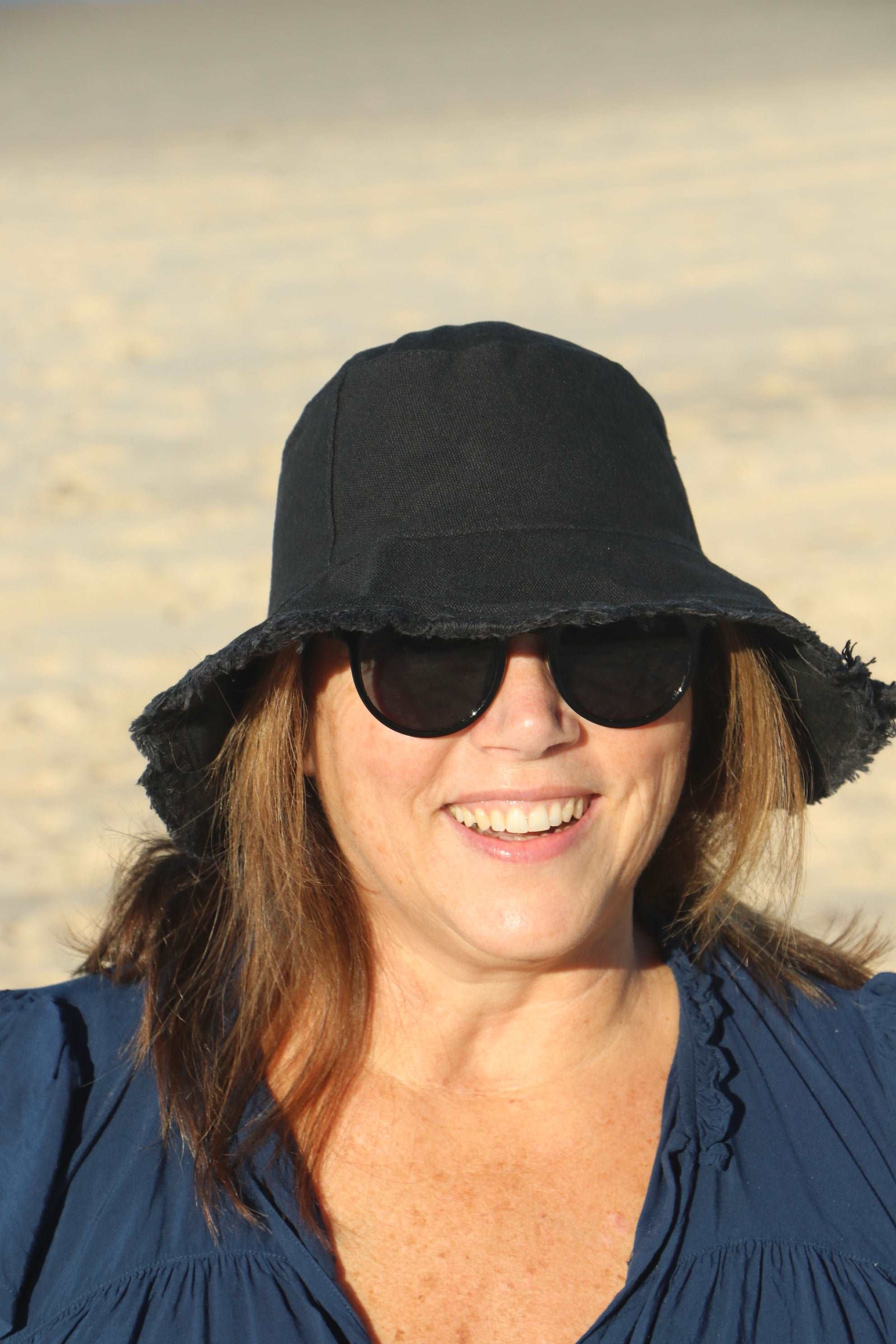 smiling lady at the beach wearing a black coloured cotton bucket hat. great sun protection. wide brim that stays in place due to wire in the rim