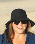 smiling lady at the beach wearing a black coloured cotton bucket hat. great sun protection. wide brim that stays in place due to wire in the rim
